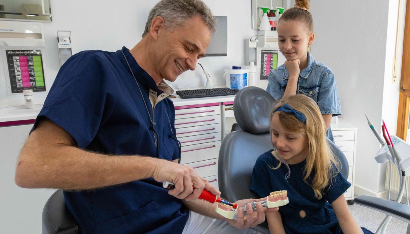 Freundlicher Zahnarzt erklärt zwei Kindern anhand von Modell, wie man richtig Zähne putzt